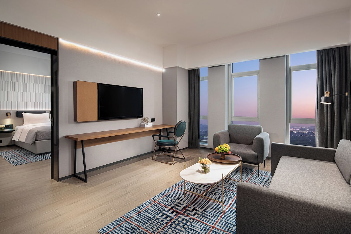 A modern hotel room with a large TV, a sofa, a chair, a small table with flowers, and a view of the city at sunset.