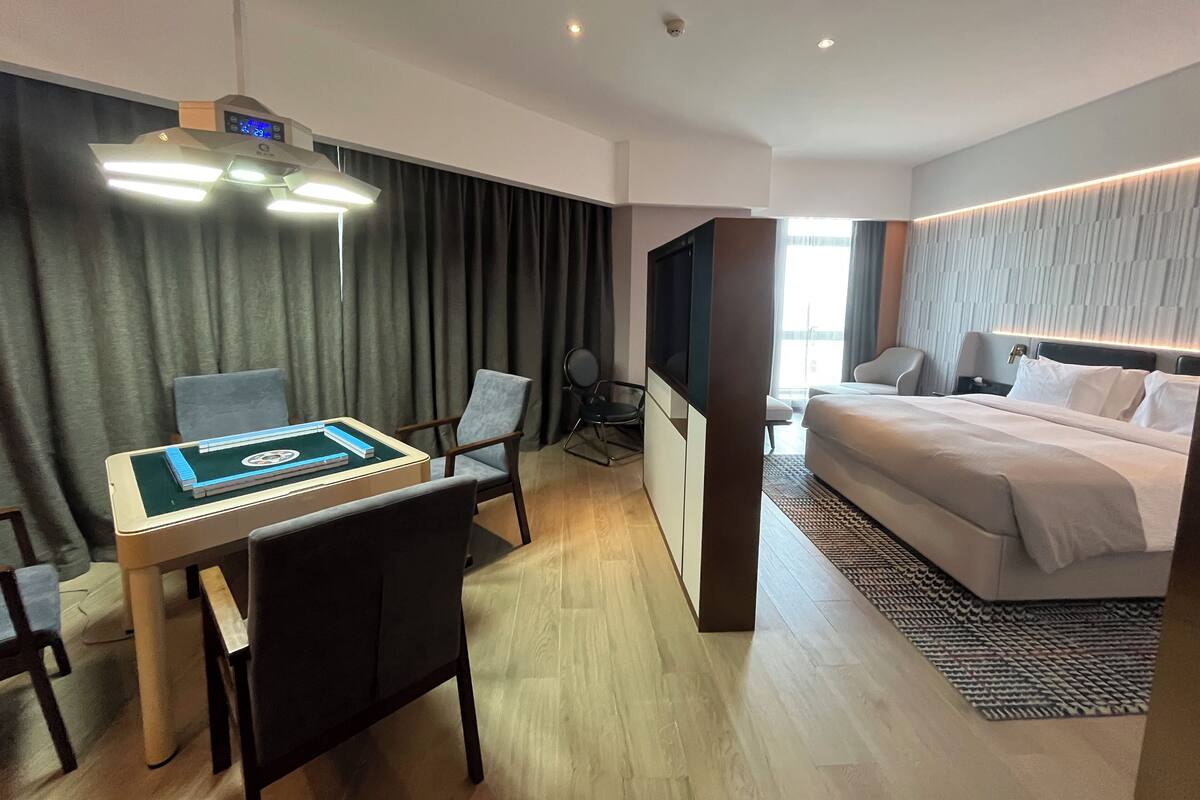 A modern hotel room with a bed, a desk with a computer, a chair, a television, and a large window.