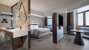 Una habitación de hotel moderna con una cama grande, un área de descanso con una mesa, un baño con tocador y un ventanal con vistas a la ciudad.