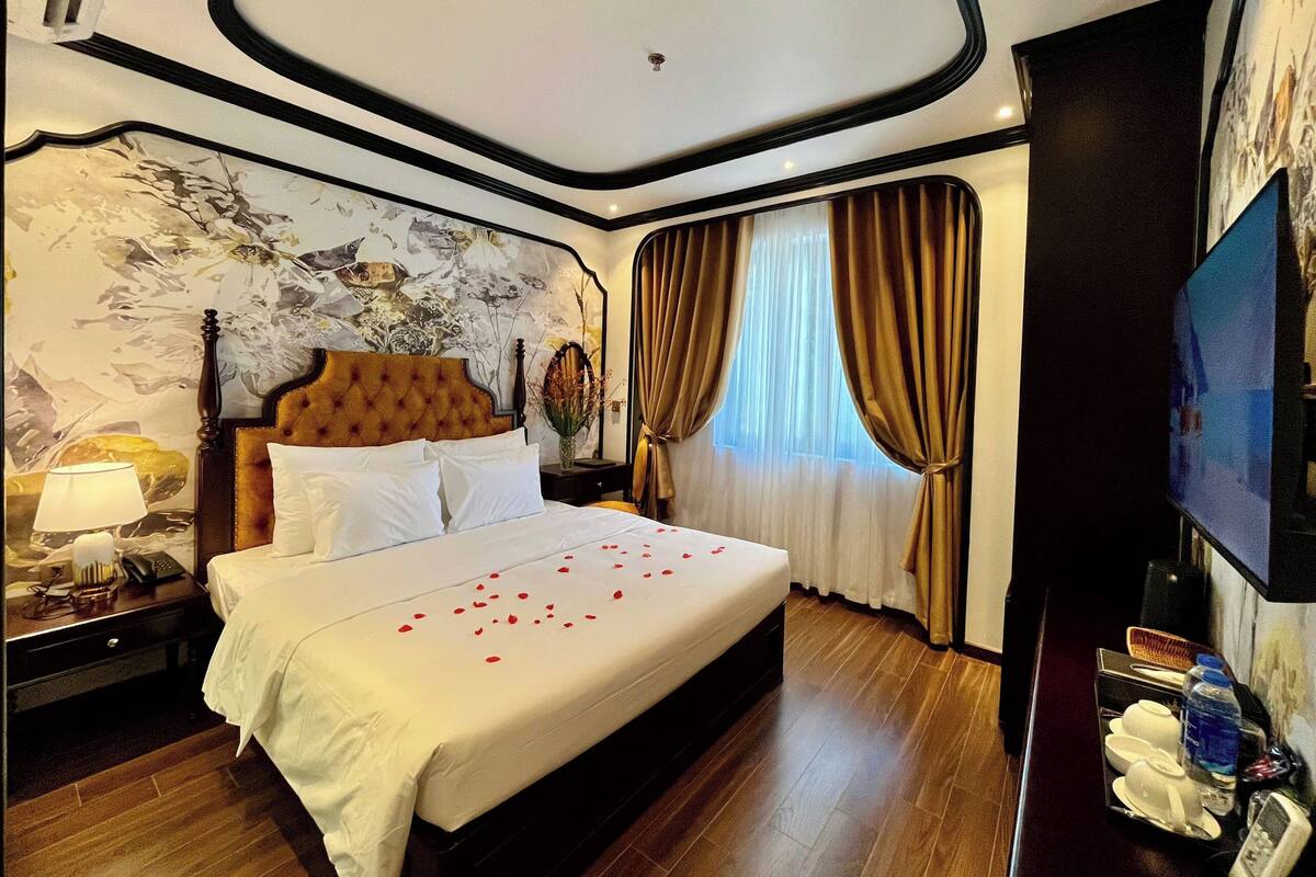 A hotel room with a large bed, a nightstand, a lamp, a vase with flowers, and a television.