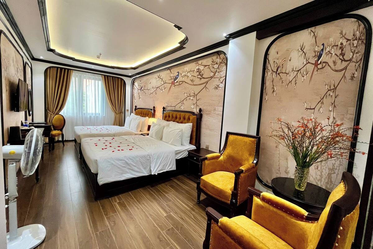 A hotel room with two beds, a sitting area with a chair, a table, and a decorative wall.