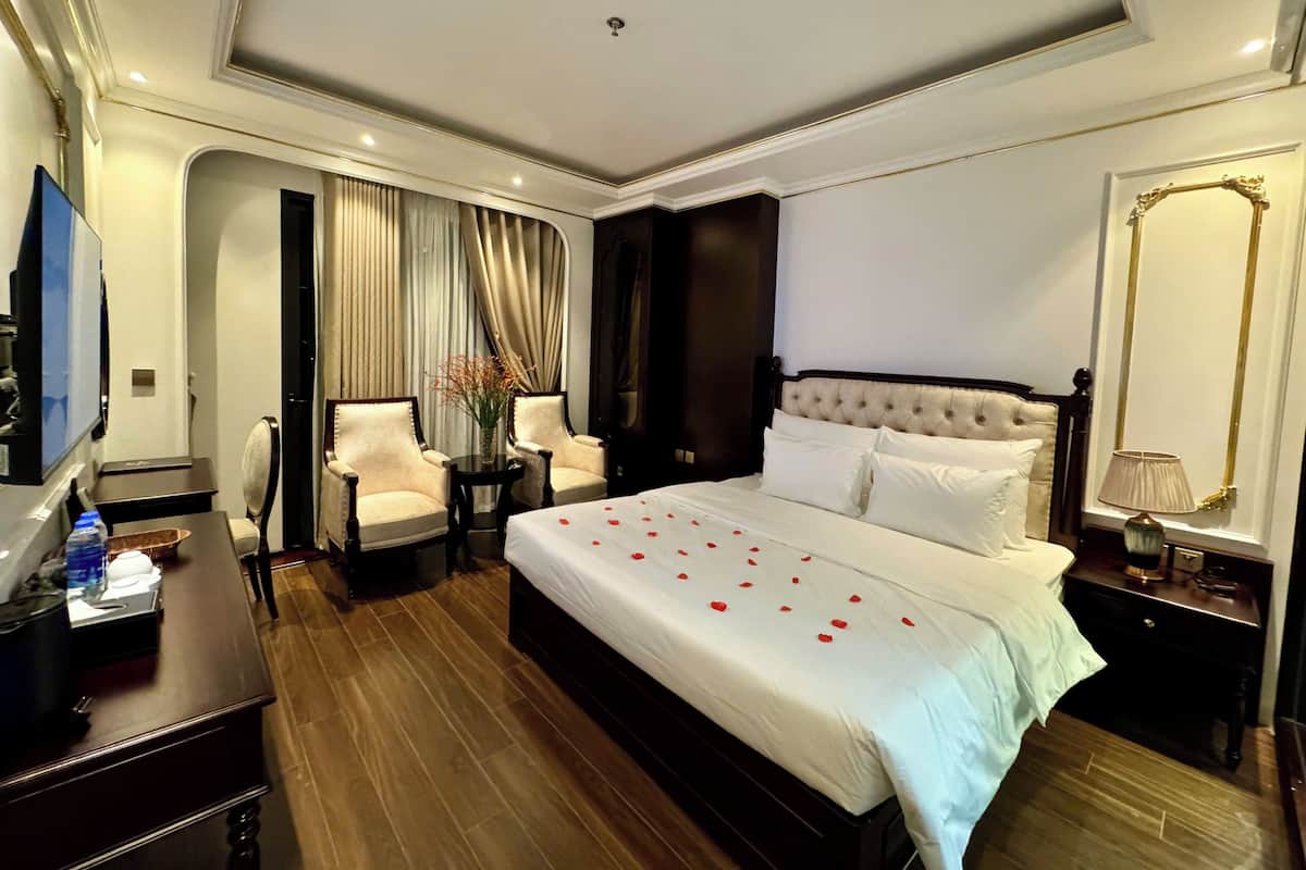 A hotel room with a large bed, two armchairs, a small table with a vase, a TV, and a nightstand with a lamp.