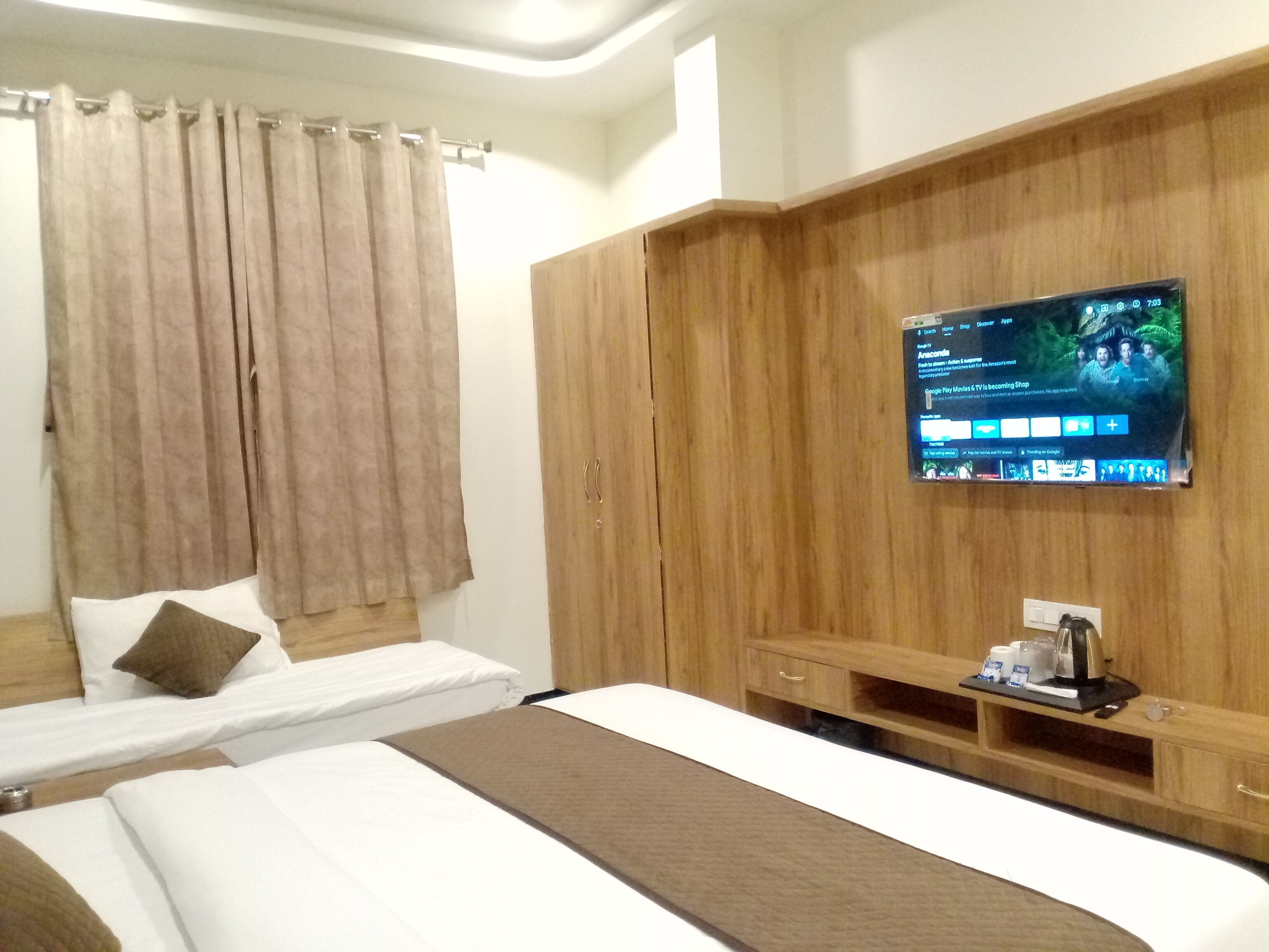 Hotellihuone, jossa on kaksi sänkyä, puinen vaatekaappi, seinälle asennettu taulutelevisio ja pieni pöytä, jossa on hygieniatarvikkeita.