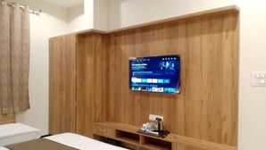Habitación de hotel con cama, televisor montado en una pared de madera, una estantería pequeña con un hervidor y una ventana con cortinas.