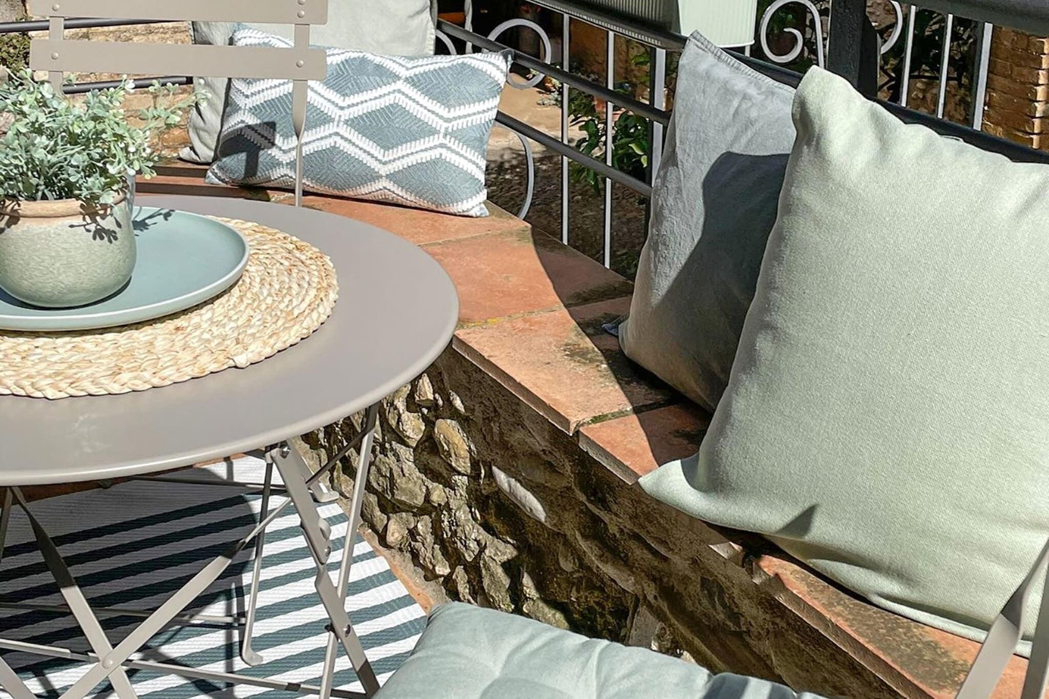 A patio with a round table, a woven placemat, and a potted plant. There are two cushioned chairs with pillows, and a stone wall behind the seating area.