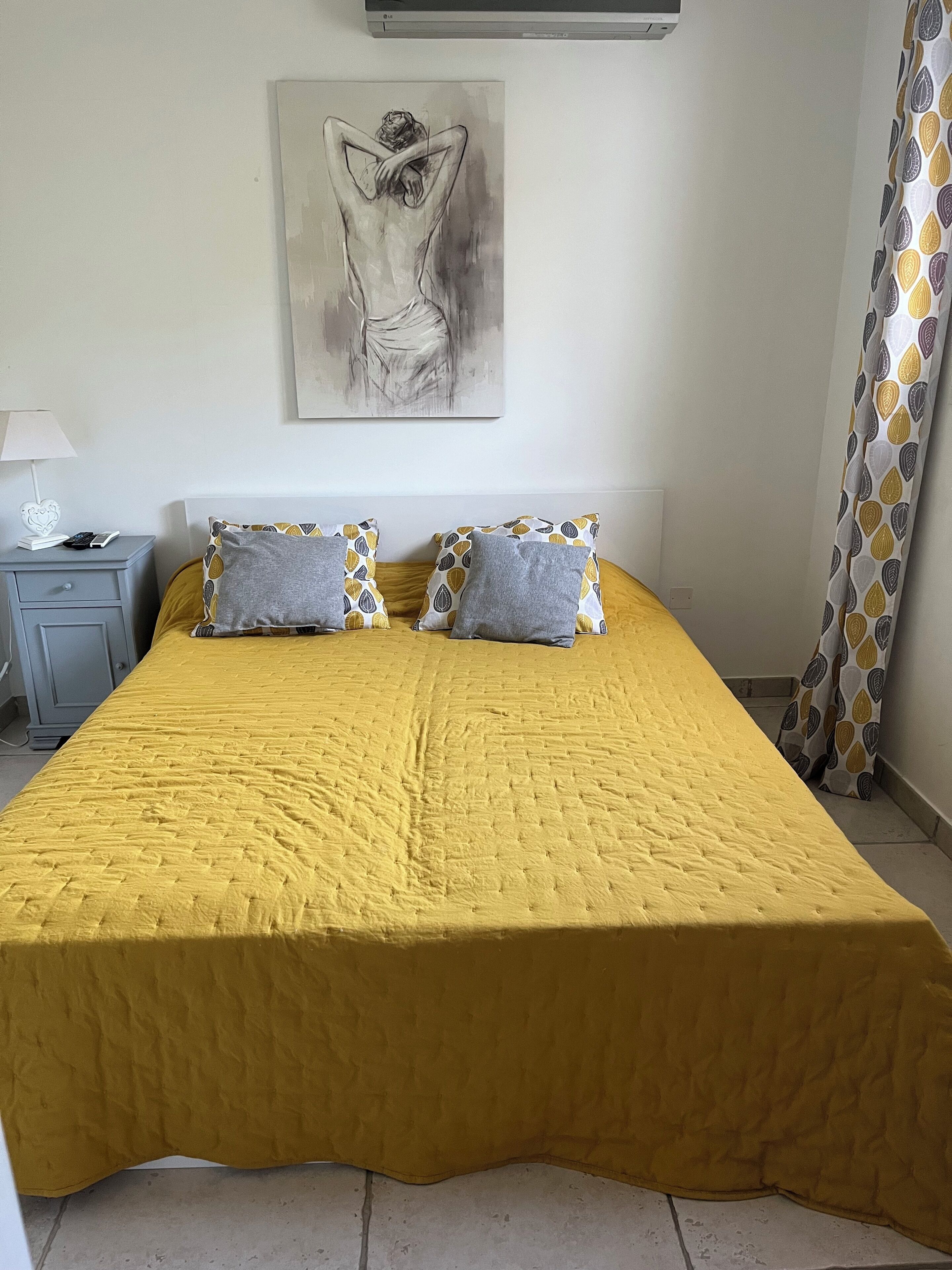 A bed with a yellow quilt, two pillows with a leaf pattern, a bedside table with a lamp, and a wall-mounted air conditioner.
