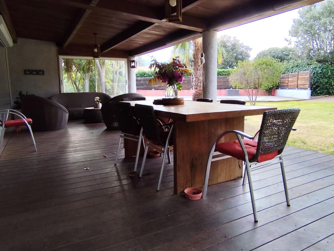 Une terrasse extérieure avec une table et des chaises en bois, un vase rempli de fleurs et une vue sur le jardin.
