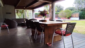 An outdoor patio with a wooden table and chairs, a vase with flowers, and a view of the garden.