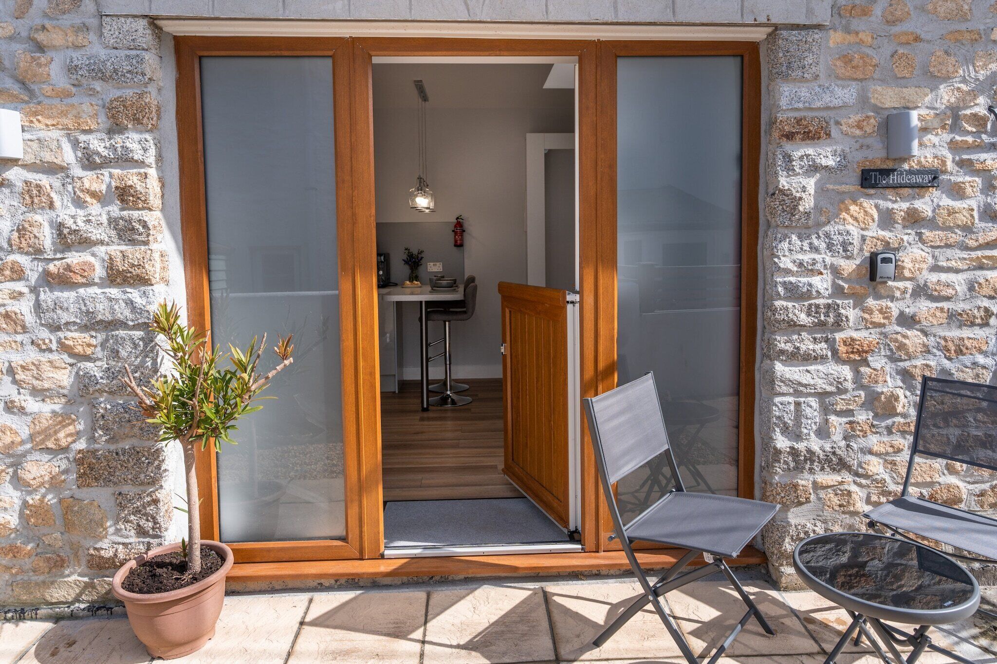 Ein Steinhaus mit einer Holztür, die zu einem Innenbereich führt. Vor der Tür steht ein Blumentopf, und auf der Terrasse sind zwei Stühle aufgestellt.