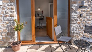 A stone building with a wooden door leading to an interior space. There is a potted plant outside, and two chairs are placed on the patio.