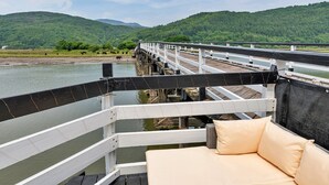 A deck with a white railing overlooking a river, a beige sofa, and a view of green hills in the distance. - Cottage by Mawddach Trail with Stunning Views (Dolgellau)