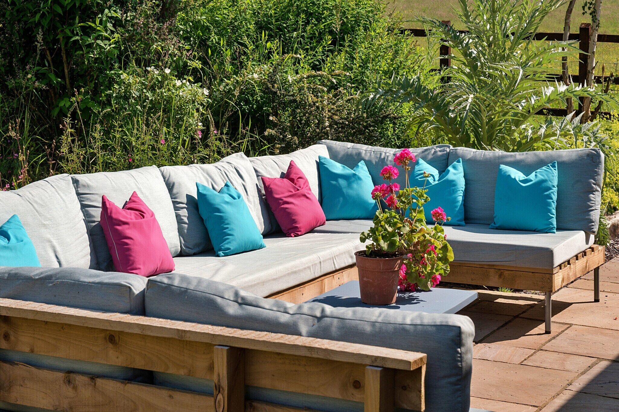 A grey sectional sofa with colorful cushions, a wooden bench, and a potted plant with pink flowers.