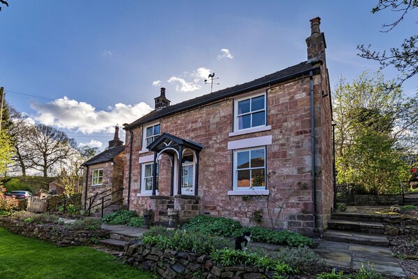 A stone house with a central entrance, a stone staircase, and a garden with a cat.