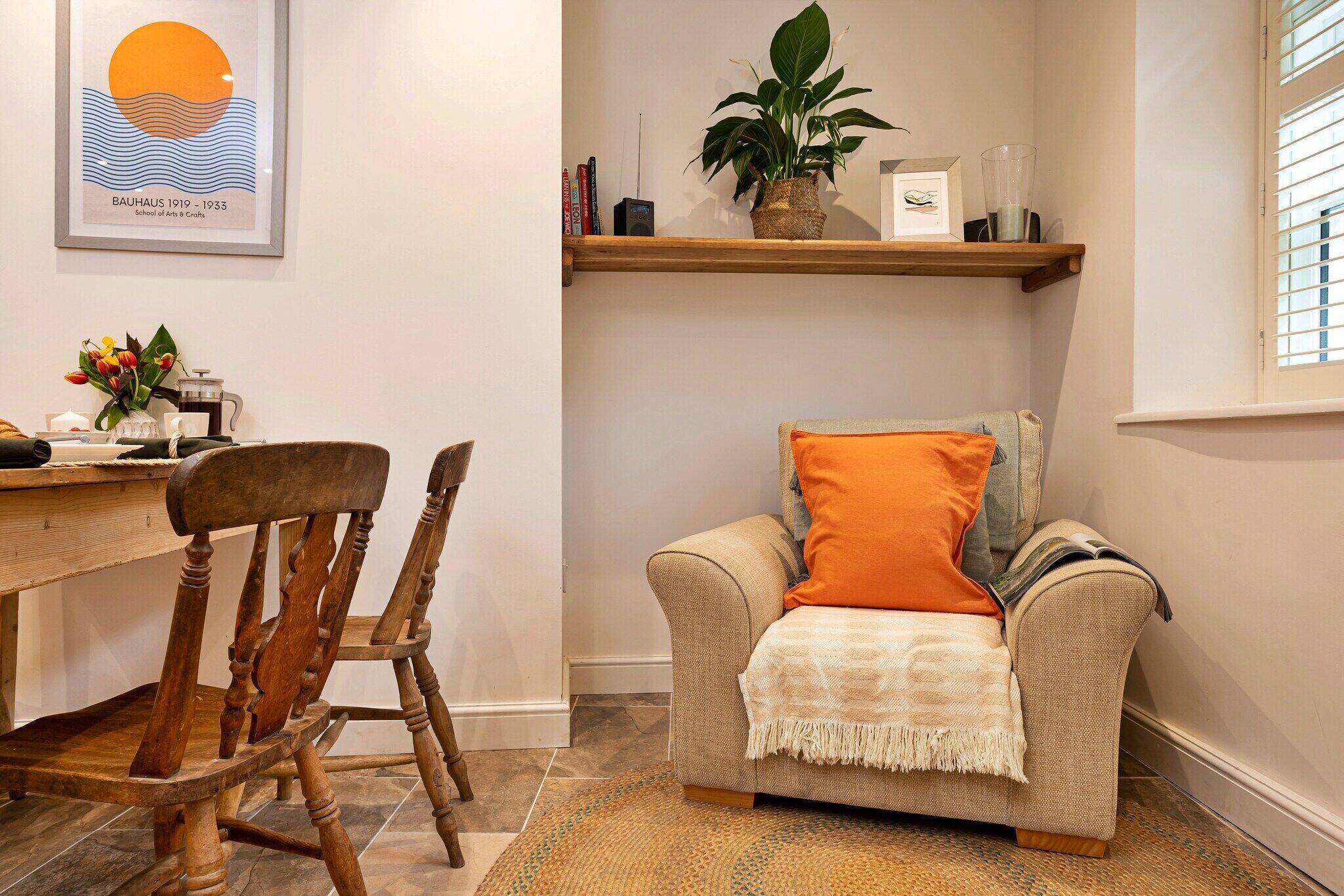 A cozy living area with a wooden chair, a beige sofa with an orange pillow, a small table with a vase of flowers, and a shelf with books and a plant.