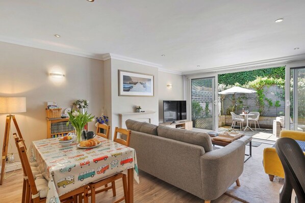 A modern living room with a sofa, dining table, and a TV. There is a balcony with outdoor furniture and a view of greenery.