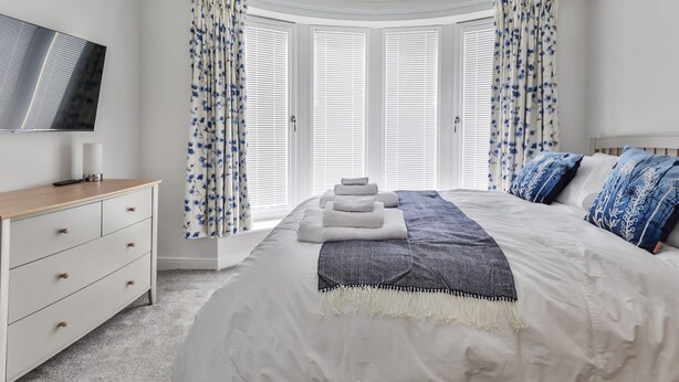 Une chambre avec un grand lit, un téléviseur fixé au mur et une vue sur l’extérieur grâce à une grande fenêtre agrémentée de rideaux.