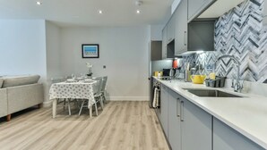 Et moderne køkken med grå skabe, en hvid bordplade og en backsplash med et fiskebensmønster. Der er et spiseområde med et bord dækket til to personer, en grå sofa og et indramet billede på væggen.