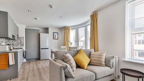 A modern living room with a grey sofa, a small table, and a kitchen area with grey cabinets and a white refrigerator. - Apartment in Filey near the Beach (Filey)