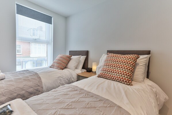 A hotel room with two beds, each with a patterned pillow, a bedside table with a lamp, and a window with blinds.