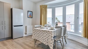 A modern kitchen with a white refrigerator, a dining table with chairs, and a bay window with curtains.