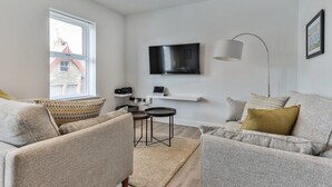 A modern living room with a sofa set, a flat-screen TV mounted on the wall, a floor lamp, and a small table with a rug.