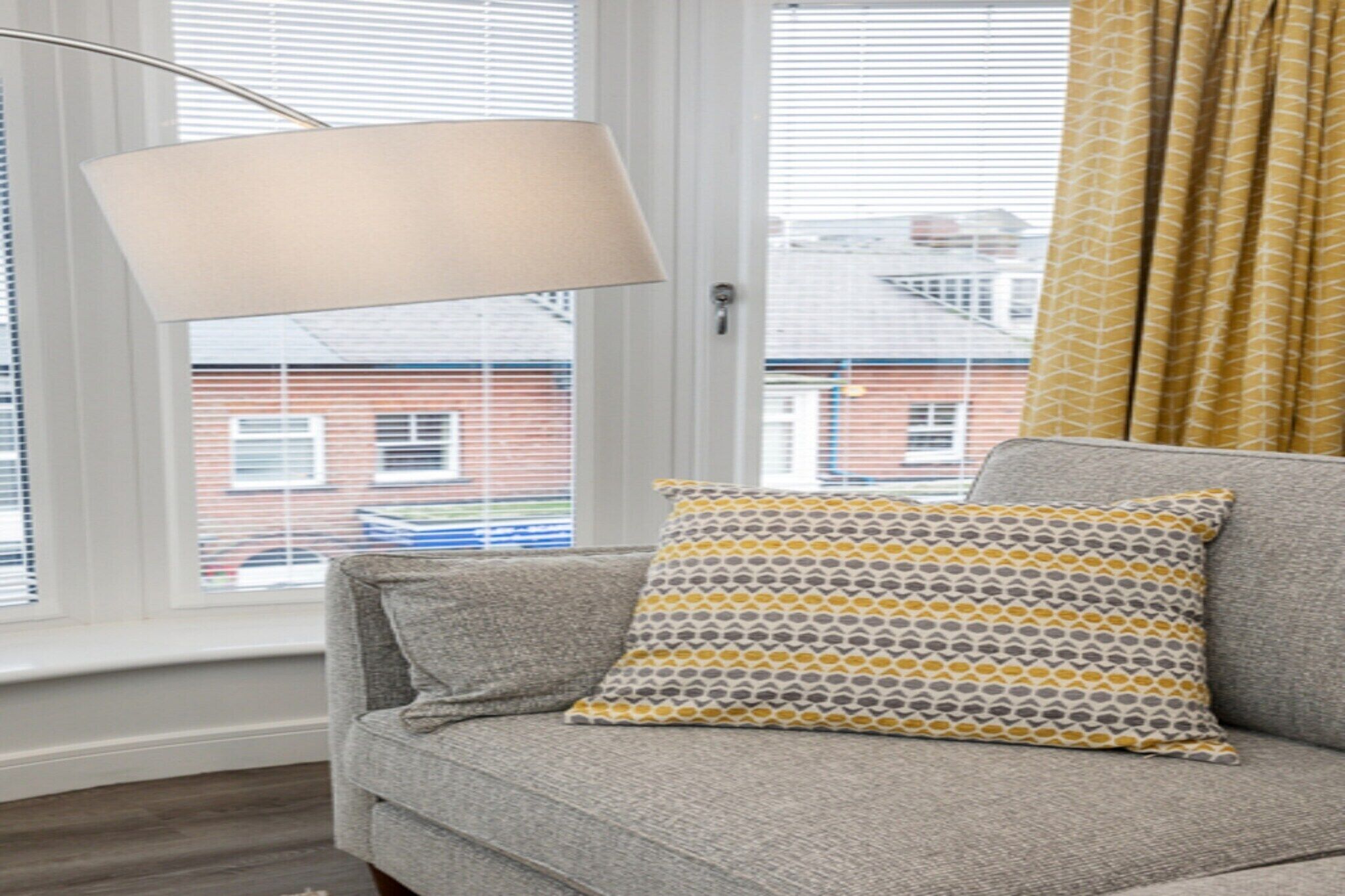 Ein Wohnzimmer mit grauem Sofa, einem gemusterten Zierkissen, einer Stehlampe und Blick auf ein Backsteingebäude durch das Fenster.