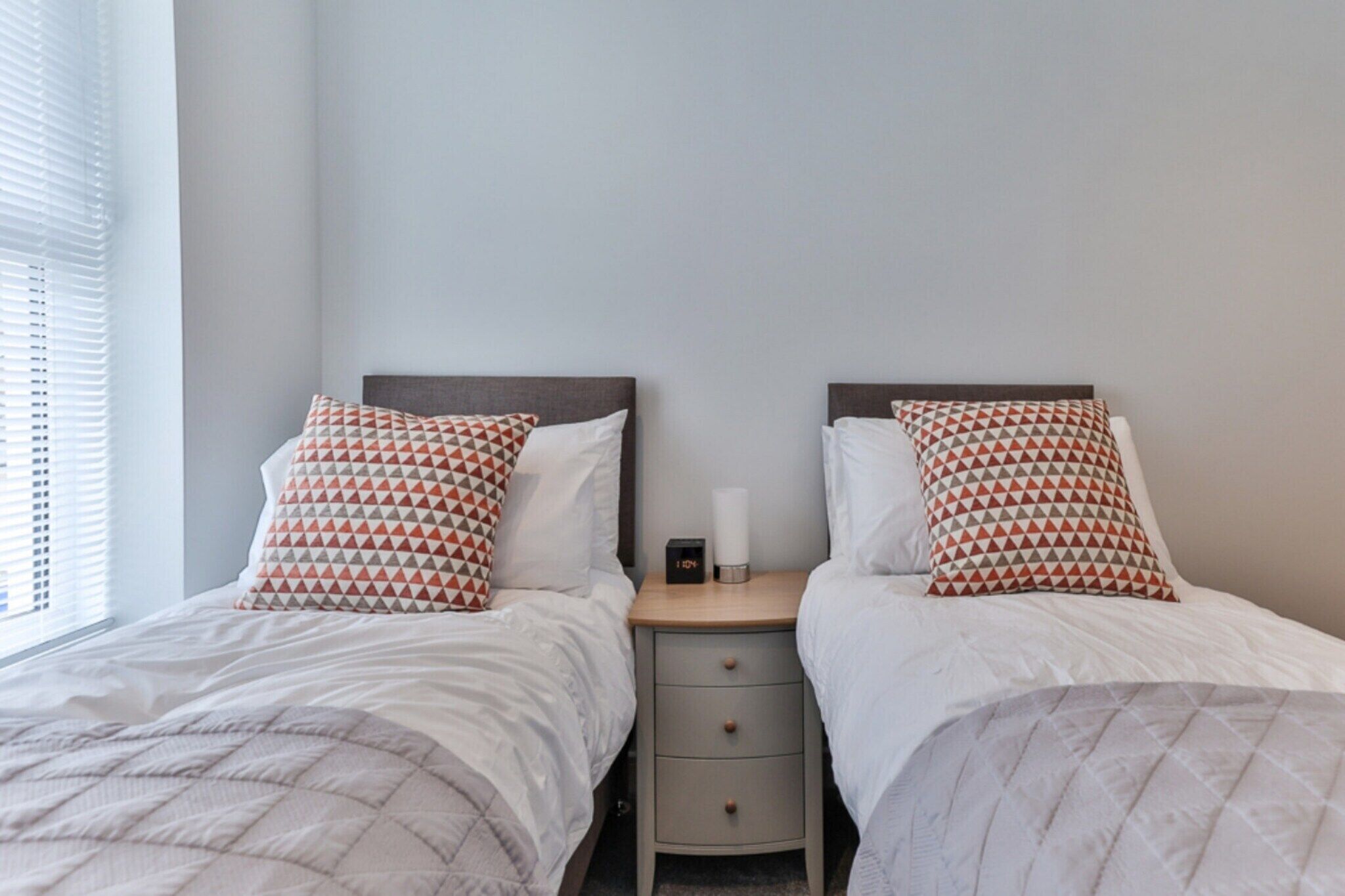 A bedroom with two beds, each with a patterned pillow, a bedside table with a clock, and a window with blinds.