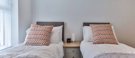 A bedroom with two beds, each with a patterned pillow, a bedside table with a clock, and a window with blinds.