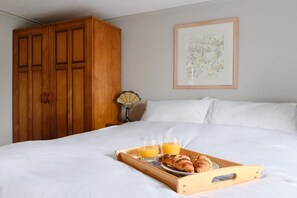 Ein ordentlich bezogenes Bett mit weißer Bettwäsche, ein Holzschrank, ein gerahmtes Blumenbild an der Wand und ein Tablett mit Croissants und Orangensaft.