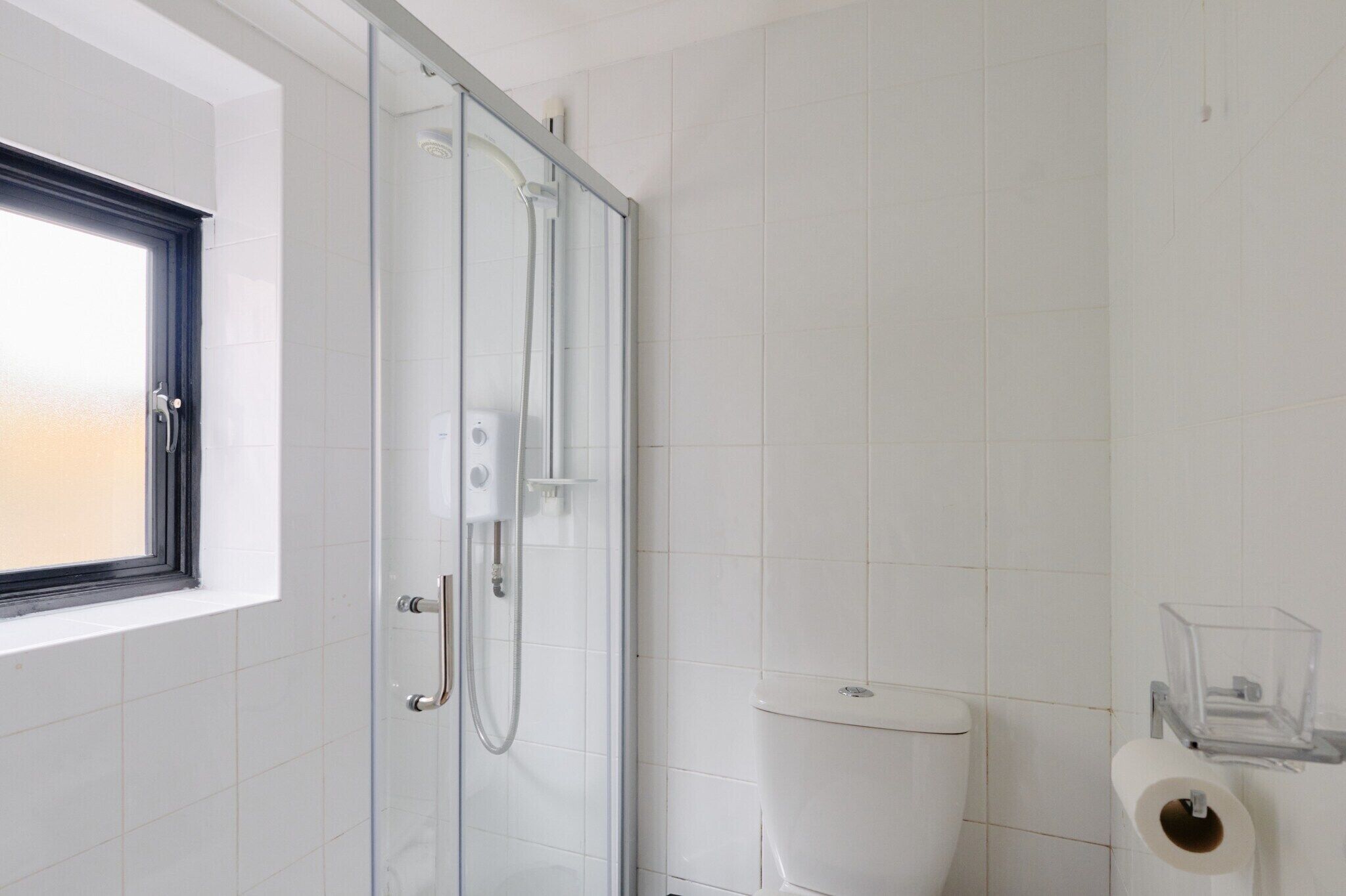 Et badeværelse med glasvæg omkring bruseniche, toilet og et vindue.