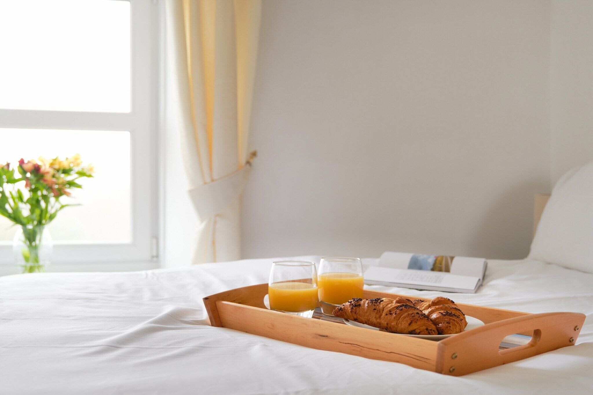 A bed with a wooden tray containing croissants and orange juice, a book, and a vase with flowers.