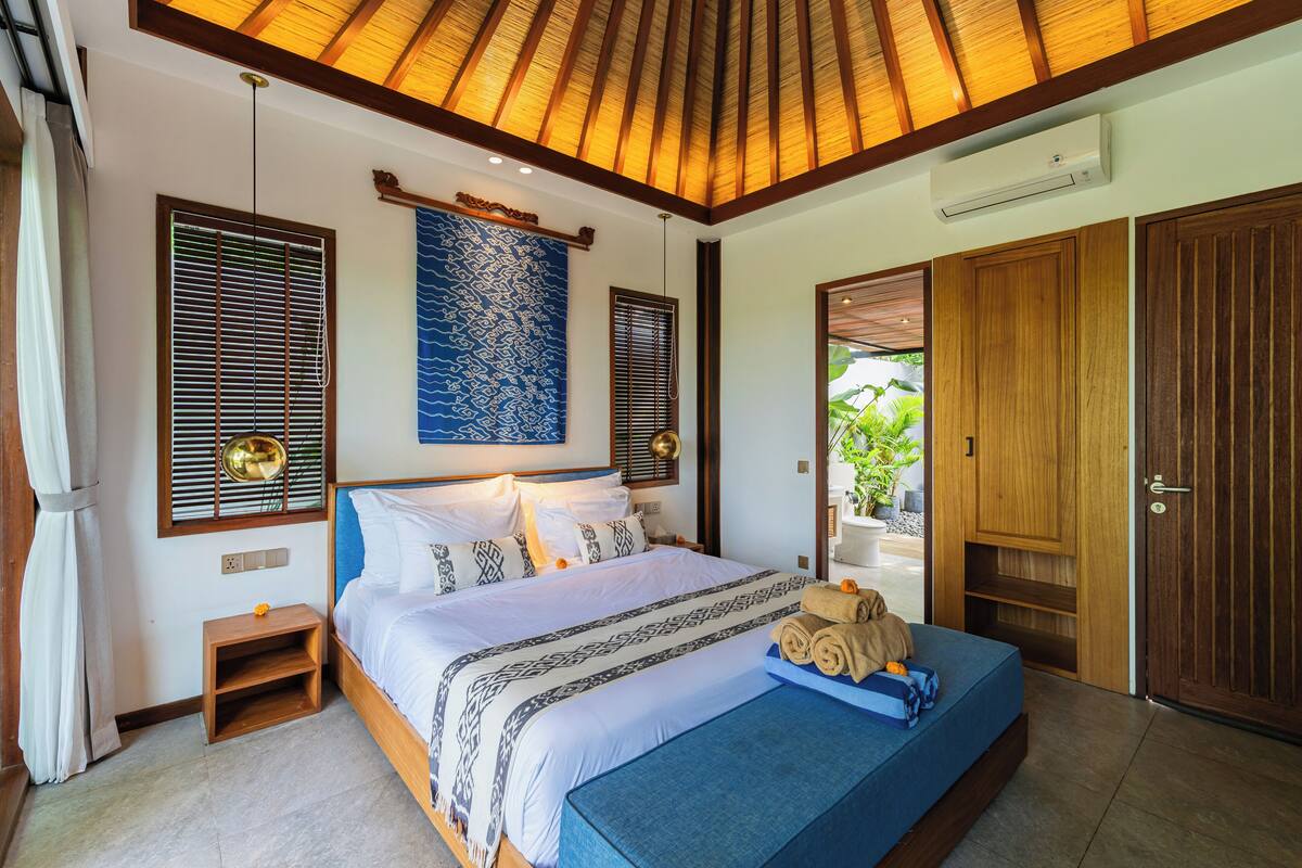 A bedroom with a large bed, wooden headboard, and a ceiling fan. There is a small wooden nightstand and a bench with towels.