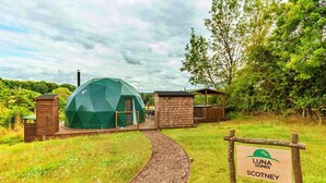 Une structure en forme de dôme géodésique, dotée d’une terrasse en bois, entourée d’une pelouse et d’arbres. Une enseigne portant le nom « Luna Domes Scotney » est visible.