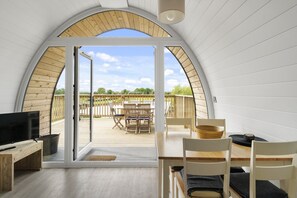 A cozy room with a large arched window, a flat-screen TV, wooden furniture, and a view of an outdoor deck with chairs and a table.