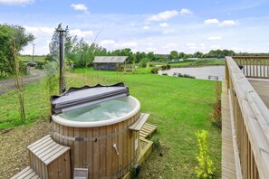 A hot tub in a grassy yard with a wooden deck and a view of a lake and distant buildings.