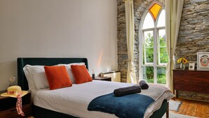 A bedroom with a stone wall, a large window with curtains, a bed with white bedding and orange pillows, a nightstand with a lamp, and a dresser with a vase of flowers. - Chapel in Snowdonia with Hot Tub & Games Room (Dinas Mawddwy)