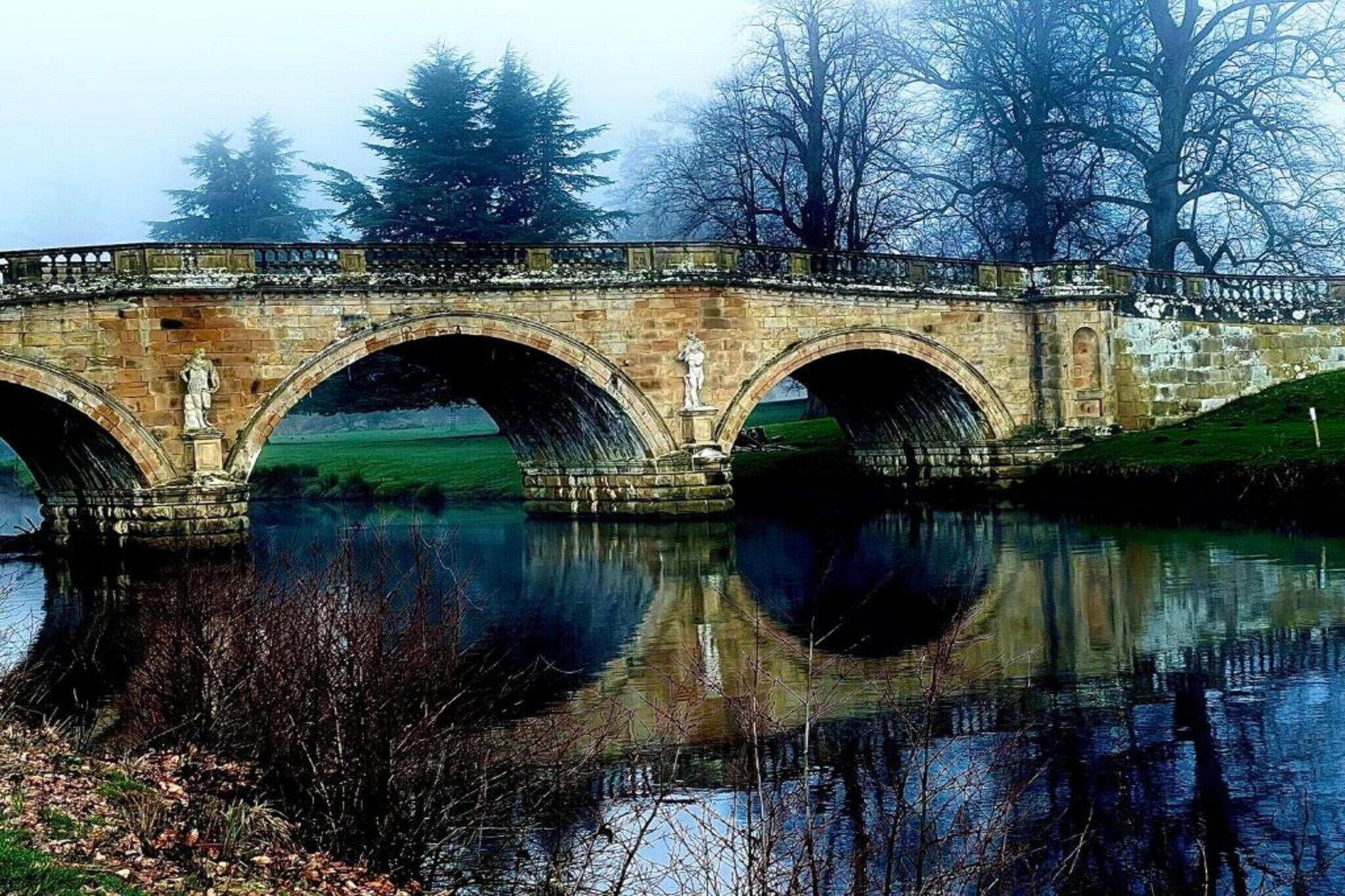A stone bridge with three arches over a calm river, surrounded by trees and grassy areas.
