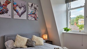 Makuuhuone, jossa on sänky, tyynyjä, yöpöytälamppu ja ikkuna, josta avautuu näkymä taloihin ja vihreään maisemaan.
