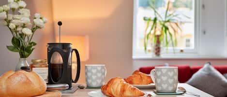 A breakfast setting with croissants, a coffee maker, and a vase of flowers on a wooden table.