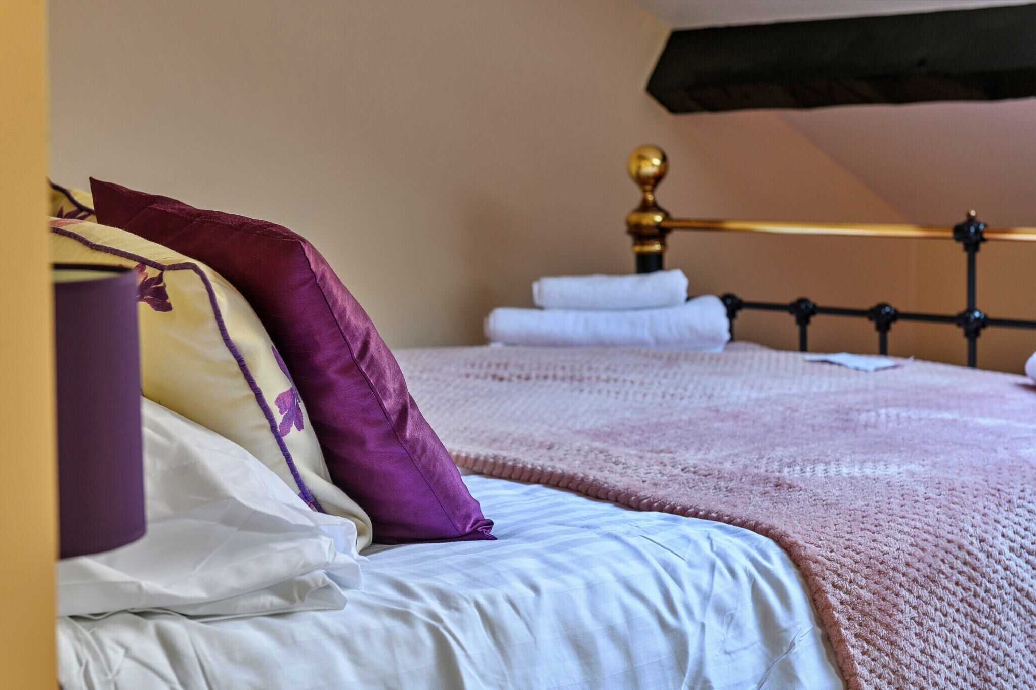 Een net opgemaakt bed met wit en paars beddengoed, een roze deken en een gouden hoofdbord.