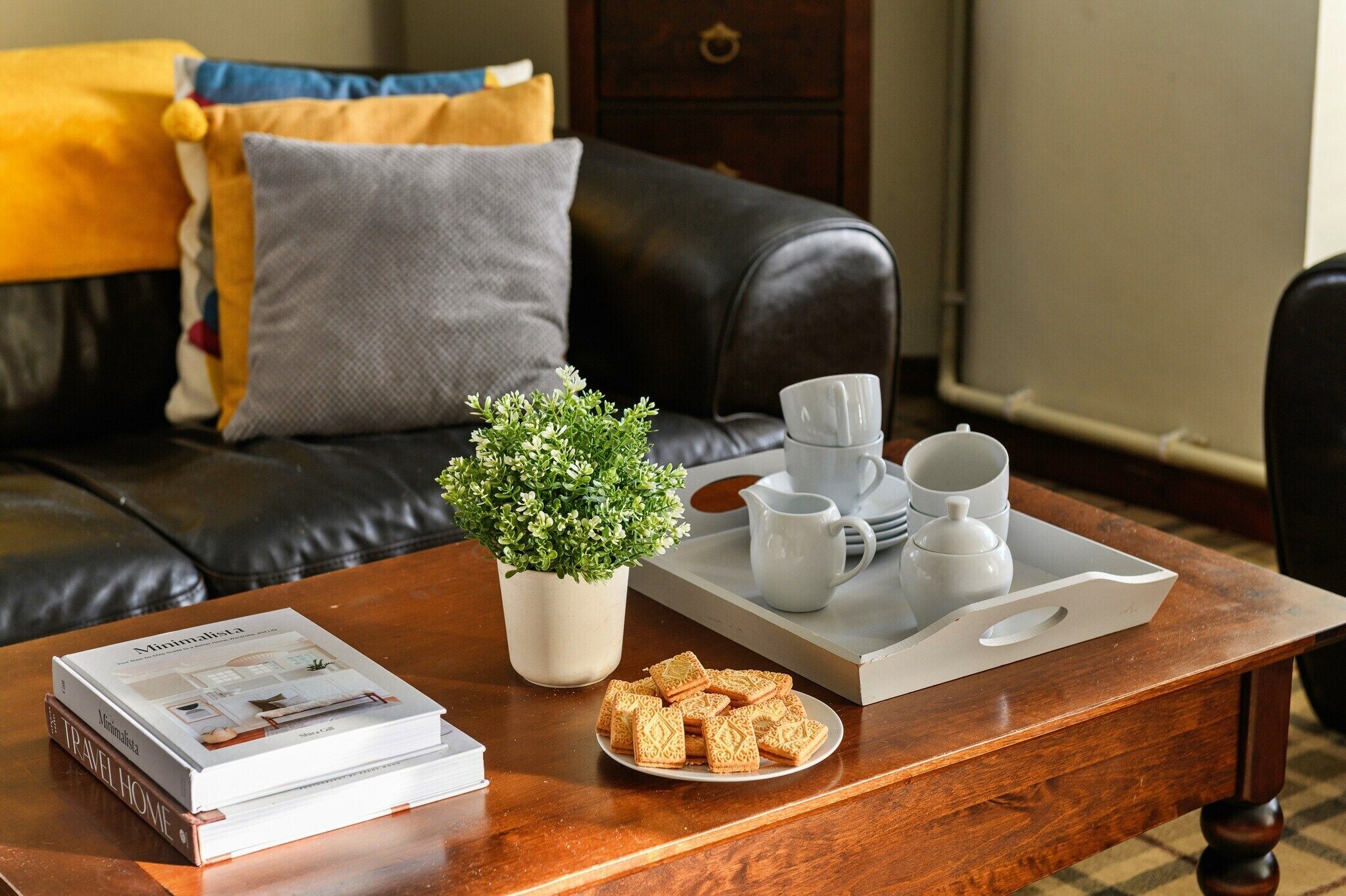 Una mesa de centro de madera con una bandeja que contiene tazas blancas, una pequeña planta en maceta y un plato de galletas. Hay libros apilados sobre la mesa, y un sofá de cuero oscuro con cojines al fondo.