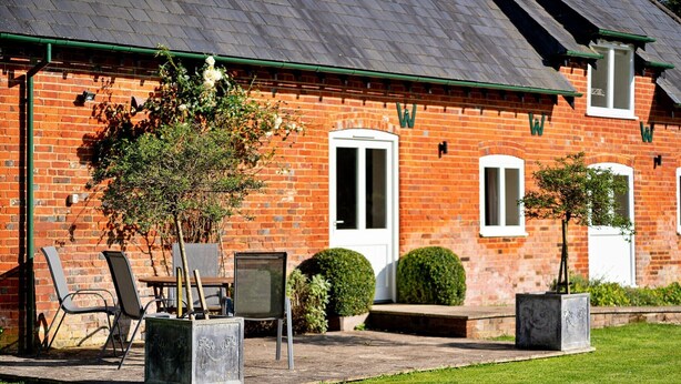 A brick building with a green roof, a white door, and a small garden area with chairs and planters. - Holiday Home in Andover near New Forest Park (Barton Stacey)