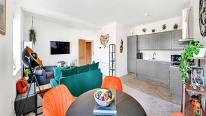 A modern living room with a round table, orange chairs, and a sofa. A kitchen with grey cabinets and a white countertop is visible in the background.