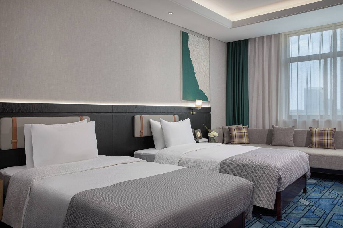 A hotel room with two beds, a sofa, a large window with curtains, and a wall-mounted artwork.