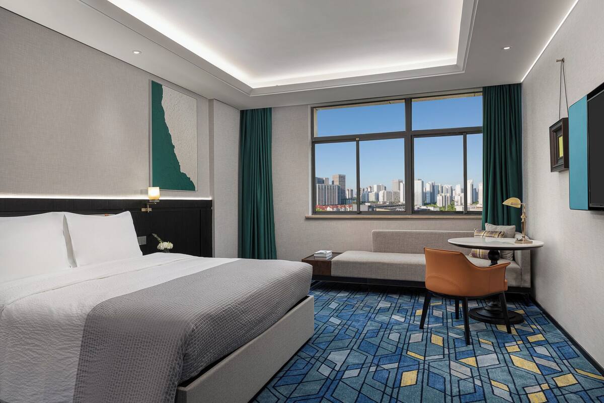A hotel room with a bed, a small sofa, a round table, and a large window offering a city view.