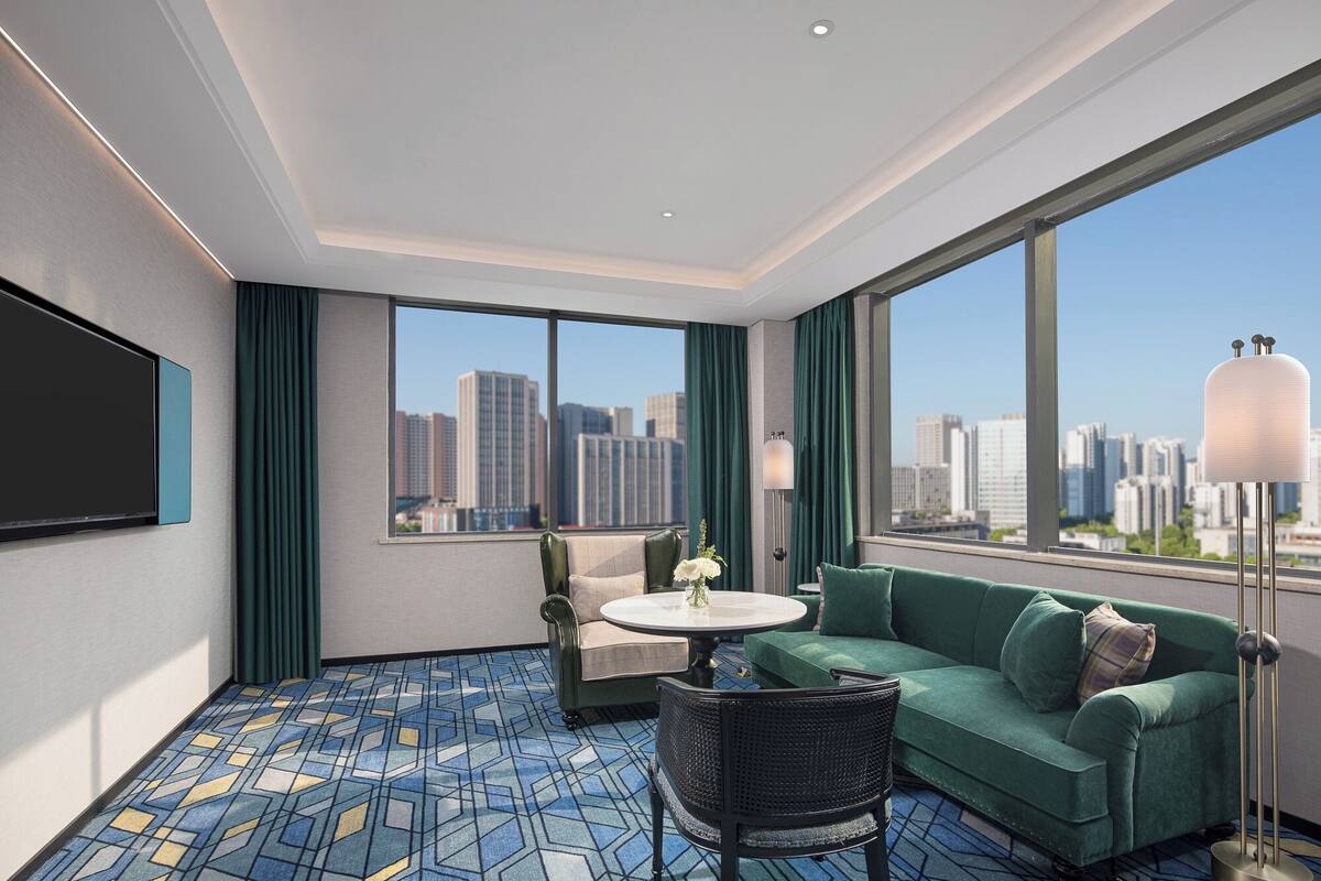 A modern hotel room with a large window offering a city view, a sofa, a chair, a small table, and a flat-screen TV.