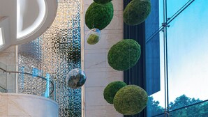 Un hall d’entrée moderne avec une grande colonne circulaire, un mur de verre orné d’un motif décoratif et plusieurs jardinières sphériques vertes.