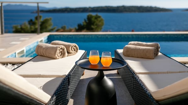 A poolside view with lounge chairs, towels, and two glasses of orange juice on a table.