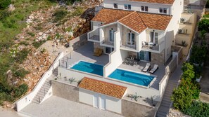A modern two-story house with a swimming pool, outdoor seating, and a stone retaining wall.