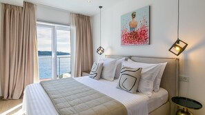 A hotel room with a bed, a painting, a nightstand, and a view of the sea.